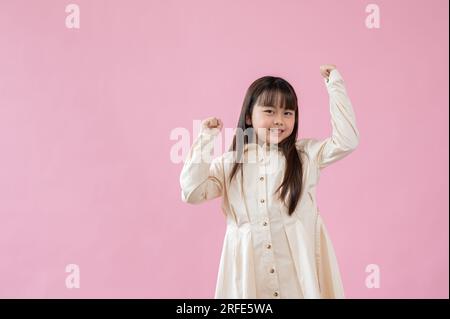 Une petite fille asiatique joyeuse et mignonne debout les mains levées, souriant à la caméra, debout sur un fond rose isolé. Banque D'Images