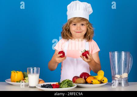 Portrait d'un enfant de 7, 8 ans dans le chapeau de cuisinier et le tablier tenir des fraises faisant la salade de fruits et la cuisson des aliments dans la cuisine. Mignon petite blonde heureuse sm Banque D'Images