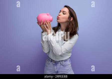 adorable femme brune de 25 ans dans une chemise et un jean décide de dépenser son épargne d'une banque de porc Banque D'Images