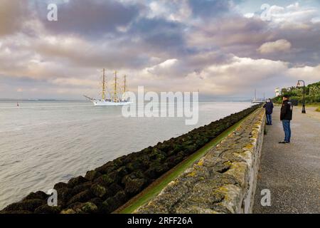 France, Calvados, Honfleur, Armada 2023, Grand Parade, Le navire-école Dar Mlodziezy navigue devant la digue où se tiennent quelques spectateurs, dans la lumière dorée de l’heure, le pont de Normandie en arrière-plan et le ciel orageux Banque D'Images