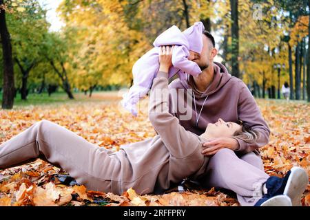 Les jeunes parents sont assis sur un plaid avec un nouveau-né dans un parc d'automne. Une mère élève un enfant dans ses bras, un père embrasse sa fille Banque D'Images