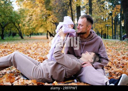 Les jeunes parents sont assis sur un plaid avec un nouveau-né dans un parc d'automne. Une mère élève un enfant dans ses bras, un père embrasse sa fille Banque D'Images