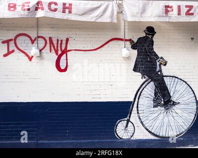 Art mural de style Banksy d'un homme sur un penny-farthing engagé dans la peinture au pistolet graffiti sur le côté d'une pizzeria de Manhattan. Banque D'Images