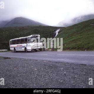 Fin des années 1970, historique, un autocar de vacances nationales stationné sur une route de montagne sur l'île de Skye, Highlands écossais, Écosse, Royaume-Uni. Un voyageur masculin devant l'autocar prenant une photo de la vallée brumeuse. National Holidays était la branche de voyage de vacances de la National bus Company, une compagnie de bus nationalisée, qui avait été créée en 1969. NBC a été rebaptisée National Express en 1974, avec une nouvelle marque de bandes bleues et blanches introduite en 1978. Au milieu des années 80, la National bus Company a été dissoute et National Holidays a été vendue à Pleasurama en 1986. Banque D'Images