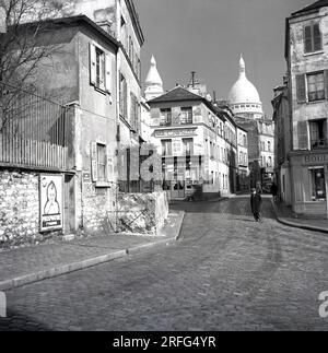 Années 1950, Paris, France, un homme marchant acorss une rue pavée dans le quartier Montmartre de la ville parisienne, célèbre pour ses artistes de rue et son église, le Sacré coeur. La photo montre les rue des Sales et le restaurant, 'le Consulatt' avec les dômes du Sacré-cœur en arrière-plan. Banque D'Images