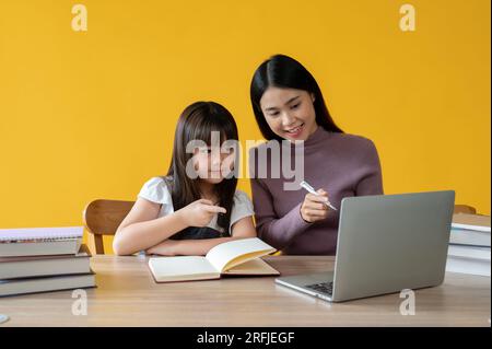 Une adorable jeune fille asiatique aime apprendre l'anglais en ligne sur un ordinateur portable avec une enseignante, assise à une table ensemble. fond jaune isolé Banque D'Images