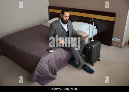 Un homme d'affaires itinérant s'assit avec un ordinateur portable sur le lit Banque D'Images