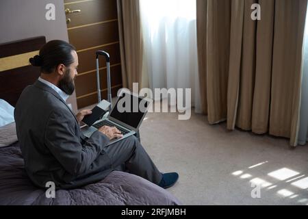 Le mâle s'assit avec un ordinateur portable sur le lit Banque D'Images