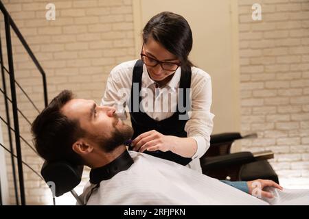 Femme barbier travaille avec la barbe d'un client, un homme dans une cape protectrice Banque D'Images