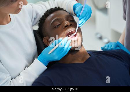 Dentiste femme concentrée vérifiant la santé des dents des clients afro-américains en utilisant le miroir buccal et la sonde de faucille, touchant chaque dent pour vérifier l'émail q Banque D'Images
