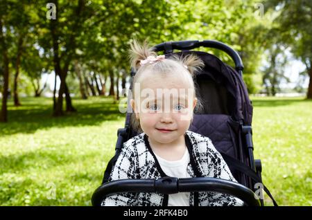 Bébé en poussette sur une promenade dans le parc d'été. Adorable petite fille assise dans une poussette, expression faciale drôle. Enfant en buggy Banque D'Images