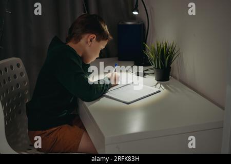 garçon caucasien faisant ses devoirs à la maison dans une pièce sombre à la lumière de la lampe. Image avec mise au point sélective Banque D'Images