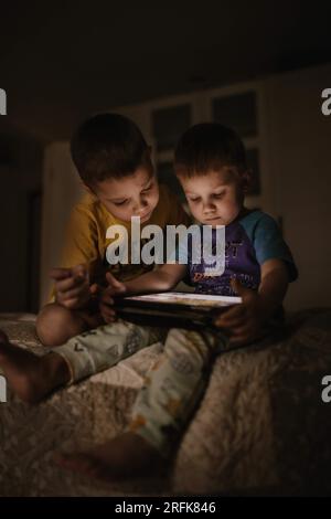 Mignon petit mignon petits garçons à l'aide de tablette jouant à des jeux vidéo ou en utilisant une application éducative, assis sur le lit dans la chambre sombre. Image avec foyer sélectif Banque D'Images