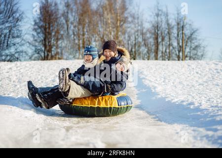 Jeune femme caucasienne jolie avec deux garçons mignons en traîneau sur la pente glacée dans le parc sur le tube de neige. Concept d'activités hivernales. Famille heureuse. Image WIT Banque D'Images