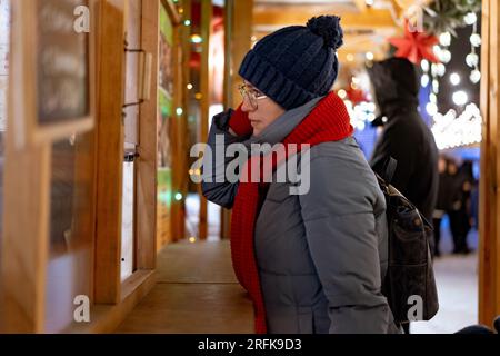 femme caucasienne portant un chapeau et une écharpe rouge allant acheter des friandises en kiosque au marché de noël. Photo de haute qualité Banque D'Images