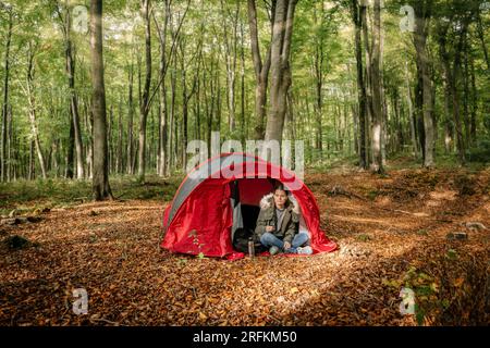 femme assise devant sa tente au milieu d'une forêt versant un café d'un thermos Banque D'Images