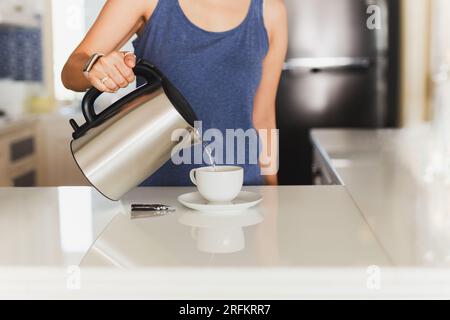 Femme versant l'eau chaude de la bouilloire dans la tasse à l'intérieur. Banque D'Images