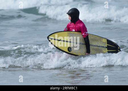 Un jeune surfeur mâle transportant son surf et marchant hors de la mer après avoir conté à la compétition de surf RIP Curl Grom Search au Fistral au ne Banque D'Images