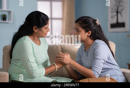 Fille indienne consolant sa mère déprimée ou triste en se tenant les mains tout en étant assise sur un canapé à la maison - concept de compassion, confort émotionnel et Banque D'Images