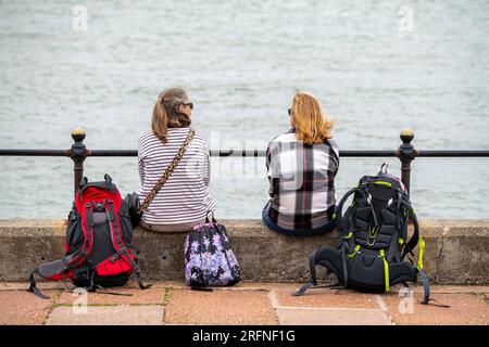 deux jeunes femmes assises sur un mur au bord de la mer avec leurs sacs à dos au sol derrière elles. deux campeurs féminins faisant une pause au bord de la mer. Banque D'Images