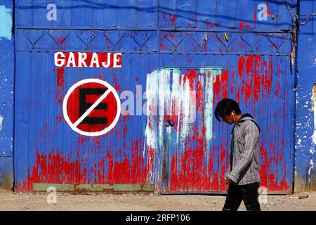 Jeune portant des écouteurs marchant devant le symbole d'absence de stationnement sur les portes de garage métalliques peintes en bleu et rouge, El Alto, Bolivie Banque D'Images