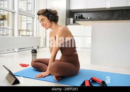 Souriant brune fille de fitness, écoute de la musique dans des écouteurs, regarde la vidéo en ligne à partir de la tablette, apprend les poses de yoga, conseils d'entraînement, fait des exercices de pilates Banque D'Images