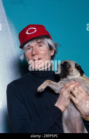 Andy Warhol pose en portant une casquette de baseball Cincinnati Reds tout en tenant un chien de race chiot en 1982. Warhol était un artiste visuel américain, réalisateur, producteur et figure de proue du mouvement pop art. Ses œuvres explorent la relation entre l'expression artistique, la publicité et la culture des célébrités qui a prospéré dans les années 1960, et couvrent une variété de médias, y compris la peinture, la sérigraphie, la photographie, le cinéma, et sculpture. Parmi ses œuvres les plus connues figurent les peintures sérigraphiées Campbell's Soup Cans et Marilyn diptyque. Photo de Bernard Gotfryd Banque D'Images