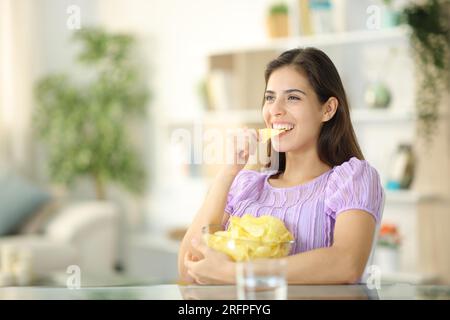 Femme heureuse mangeant des chips assis dans le salon à la maison Banque D'Images