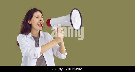 Portrait panoramique d'une belle jeune femme brune heureuse vêtue d'une chemise blanche, criant dans un mégaphone avec sa bouche grande ouverte. Banque D'Images