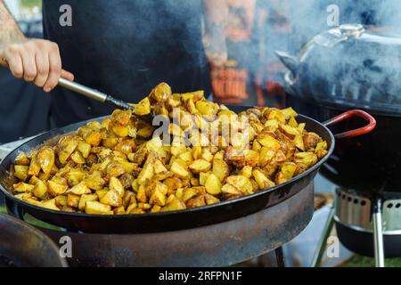 Le cuisinier mélange les pommes de terre frites en quartiers dans une grande casserole sur le gril dans la cour Banque D'Images