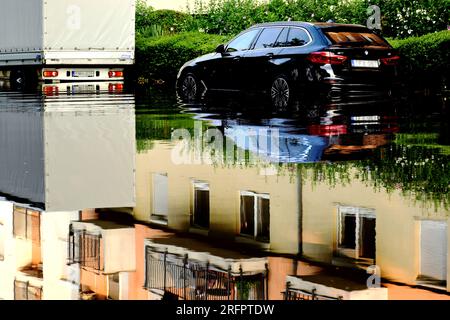 scène de rue urbaine après une forte tempête. route inondée. voiture partiellement dans l'eau de pluie profonde. pluie torrentielle. de fortes réflexions. trottoir et haie verte Banque D'Images