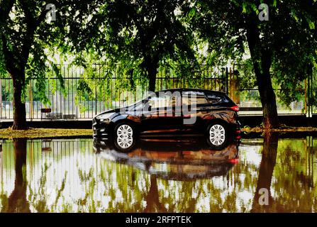 scène de rue urbaine après une forte tempête. route inondée. voiture partiellement dans l'eau de pluie profonde. pluie torrentielle. de fortes réflexions. trottoir et arbres verts Banque D'Images