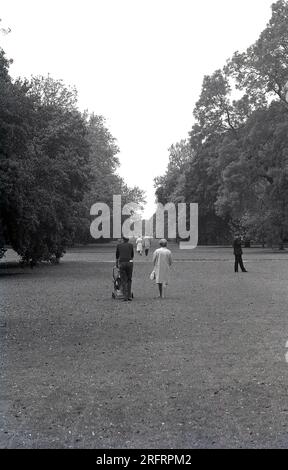 1970, historique, un couple marchant sur l'herbe dans un parc, l'homme poussant un enfant dans une petite poussette à roues, Angleterre, Royaume-Uni. Banque D'Images