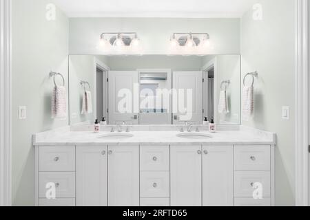 Une salle de bains avec un meuble blanc et un comptoir en marbre, murs vert clair, et une douche avec des carreaux blancs et bleus vus à travers le miroir. Banque D'Images