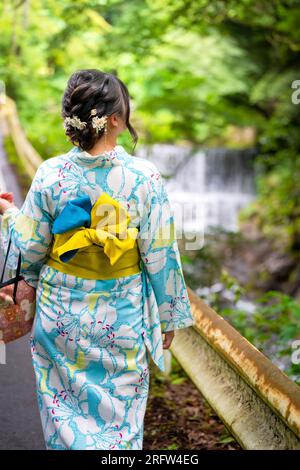 Vue arrière d'une femme portant le kimono d'été japonais yukata marchant sur la route à côté de la cascade de la forêt dans la nature. Kyoto, Japon. Banque D'Images