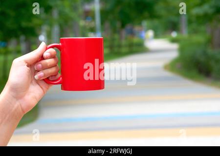 Tasse en céramique rouge dans une main féminine sur une allée verte dans le parc Banque D'Images