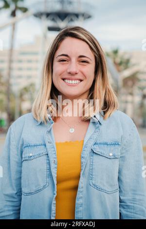 Portrait vertical de jeune femme adulte blonde heureuse debout dehors avec une expression amicale et optimiste. Femme de beauté caucasienne avec des dents blanches parfaites souriant et regardant satisfed à la caméra. Photo de haute qualité Banque D'Images