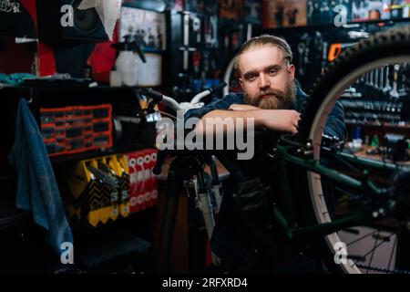 Portrait moyen de tir de beau réparateur de cyclisme avec la barbe se tenant appuyé sur le vélo dans l'atelier de réparation de vélo avec intérieur sombre, regardant Banque D'Images