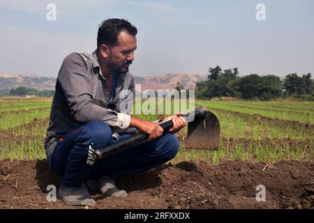 Fermier indien assis dans le champ avec une pelle, lent après avoir travaillé dans le champ, la verdure et le fond du ciel Banque D'Images