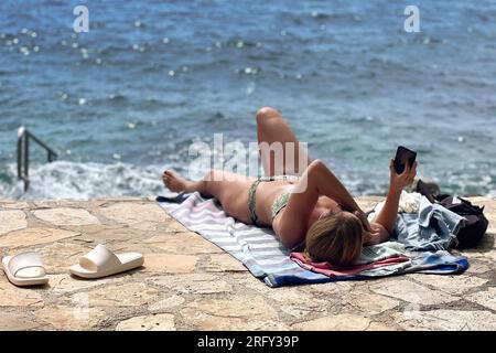 Plava Laguna, côte Adriatique près de Porec/Croatie, plage. Vacancier, jeune femme, célibataire regarde son smartphone, téléphone portable, bains de soleil sur la plage de pierre, plage rocheuse. ? Banque D'Images