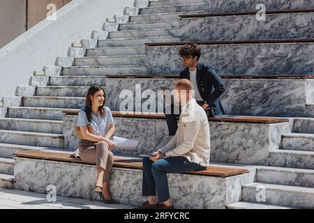 Heureux gens d'affaires assis sur les escaliers dans la zone urbaine ayant des conversations aléatoires et souriant tout en travaillant ensemble à distance Banque D'Images