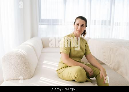 Portrait de thérapeute masseuse féminine attrayante en uniforme assis sur le canapé regardant la caméra avec expression amicale, sur le fond de Banque D'Images