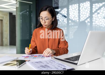 Sérieuse pensée femme financière sur le travail de papier, asiatique concentré écrire des données dans des formulaires, utiliser un ordinateur portable au travail, examiner les rapports financiers des contrats, comptable sur le lieu de travail. Banque D'Images