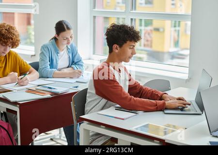 Écolier adolescent utilisant un ordinateur portable près de cahiers et camarades de classe floues dans la salle de classe à l'école Banque D'Images