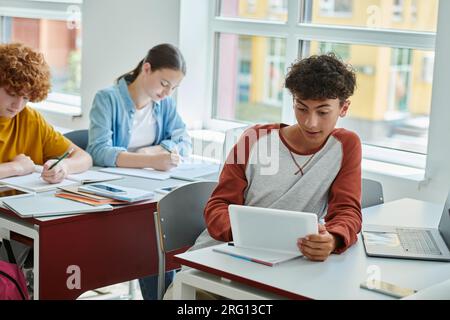 Adolescent écolier à l'aide de la tablette numérique près des appareils avec écran blanc et camarades de classe dans la salle de classe Banque D'Images