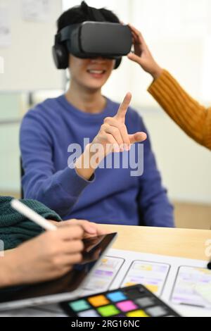 Jeune homme créatif portant un casque VR ayant une expérience de réalité virtuelle, équipe de démarrage discutant de nouveaux projets numériques à la réunion. Banque D'Images