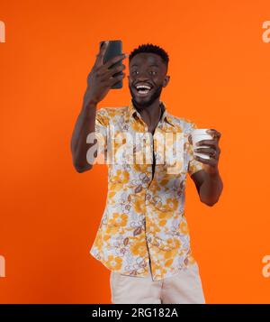 Joyeux homme afro-américain tenant smartphone et prenant l'autoportrait tout en tenant le verre de carton sur fond orange Banque D'Images
