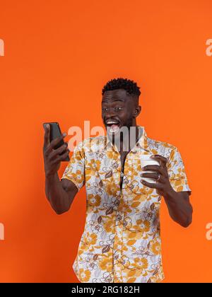 Joyeux homme afro-américain tenant smartphone et prenant l'autoportrait tout en tenant le verre de carton sur fond orange Banque D'Images