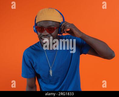 Joyeux hipster barbu élégant dans des lunettes de soleil avec chaîne de cou regardant la caméra tout en ajustant le casque et en écoutant de la musique contre le backgro orange Banque D'Images