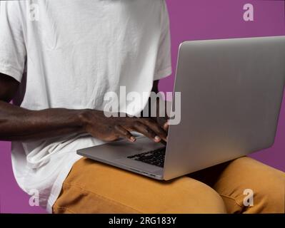 Anonyme jeune indépendant ethnique masculin dans des vêtements décontractés tout en étant assis et tapant sur le clavier de l'ordinateur portable pendant le travail pour la société à distance contre violet Banque D'Images